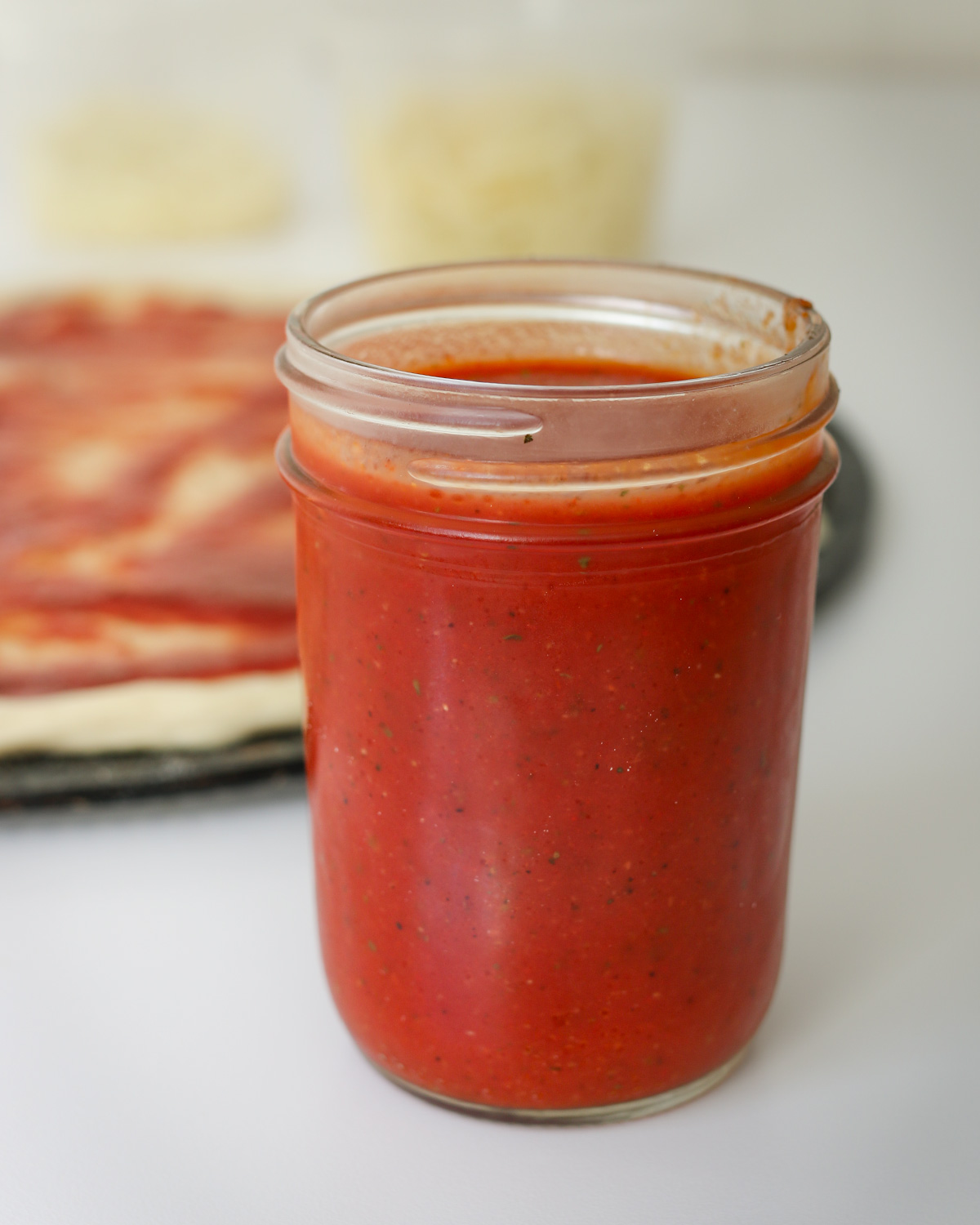 jar of pizza sauce made with tomato paste on table next to pizza and container of cheese.