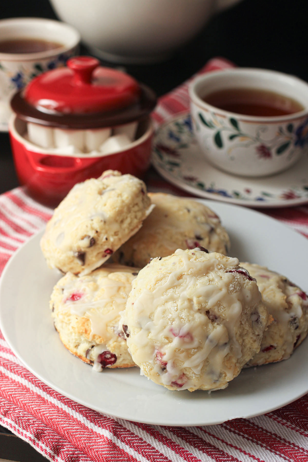 chocolate chip scones stacked on a white plate on a table with tea cups and sugar bowl.