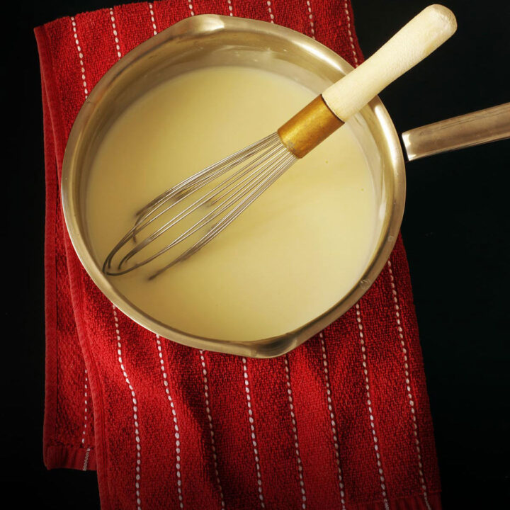 saucepan of white sauce on a red striped towel with a whisk.