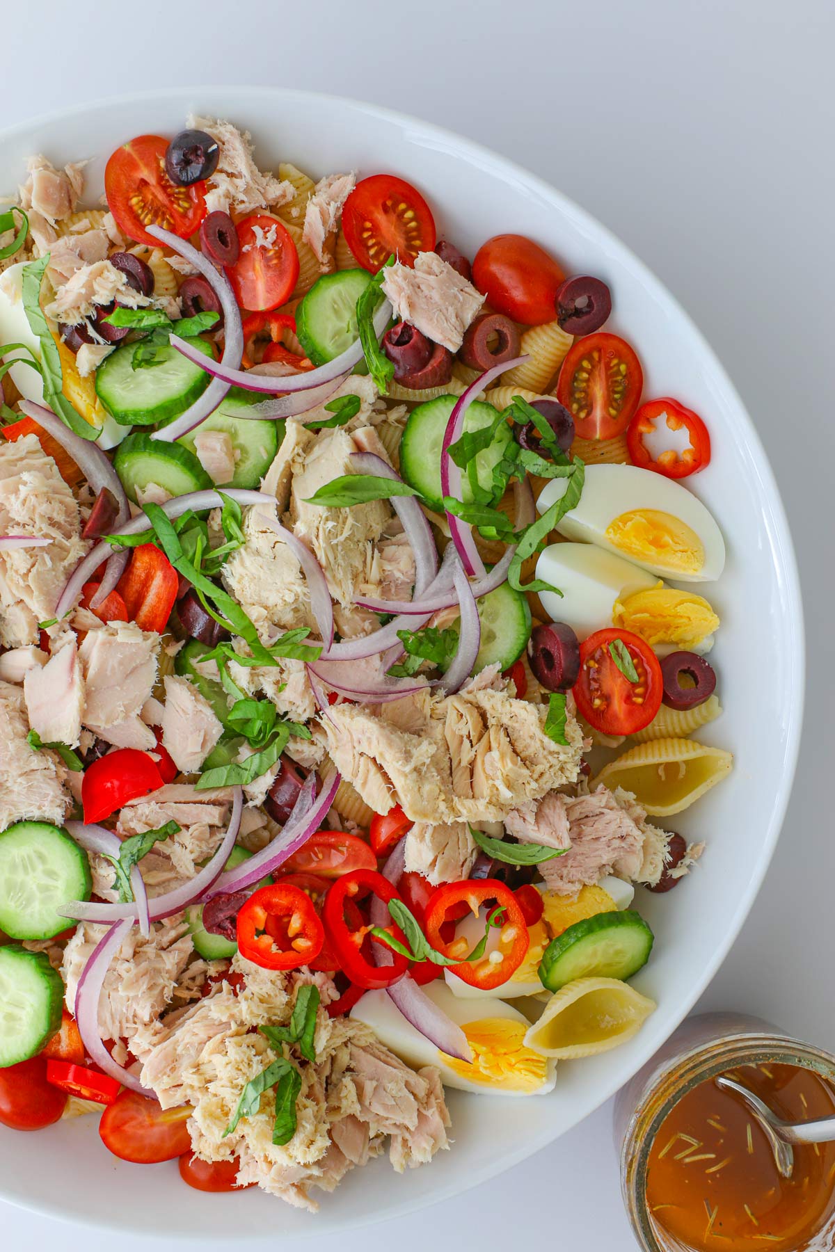 overhead shot of tuna pasta salad in white bowl on white table with jar of dressing nearby.