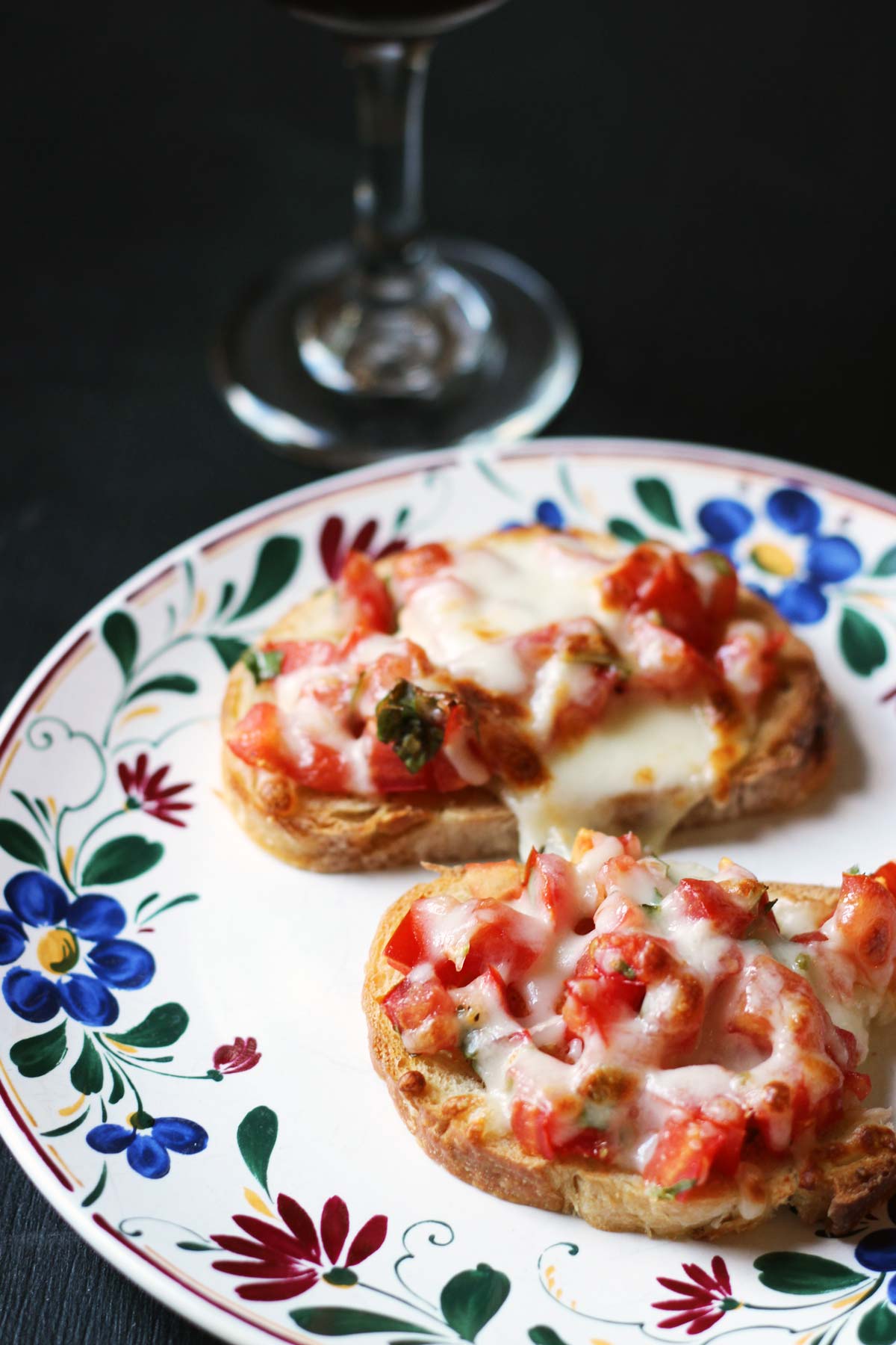 two cheese topped bruschetta on plate next to a glass of wine.
