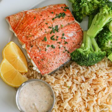 overhead shot of salmon, broccoli, and lemon wedges on a plate next to rice pilaf and tartar sauce cup.