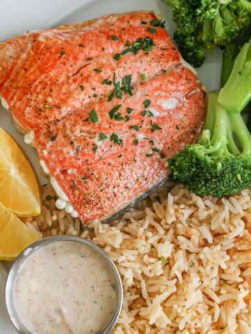 overhead shot of salmon, broccoli, and lemon wedges on a plate next to rice pilaf and tartar sauce cup.