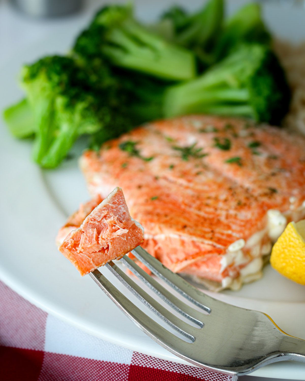 bite of fish on a fork in front of dinner plate with salmon and broccoli.