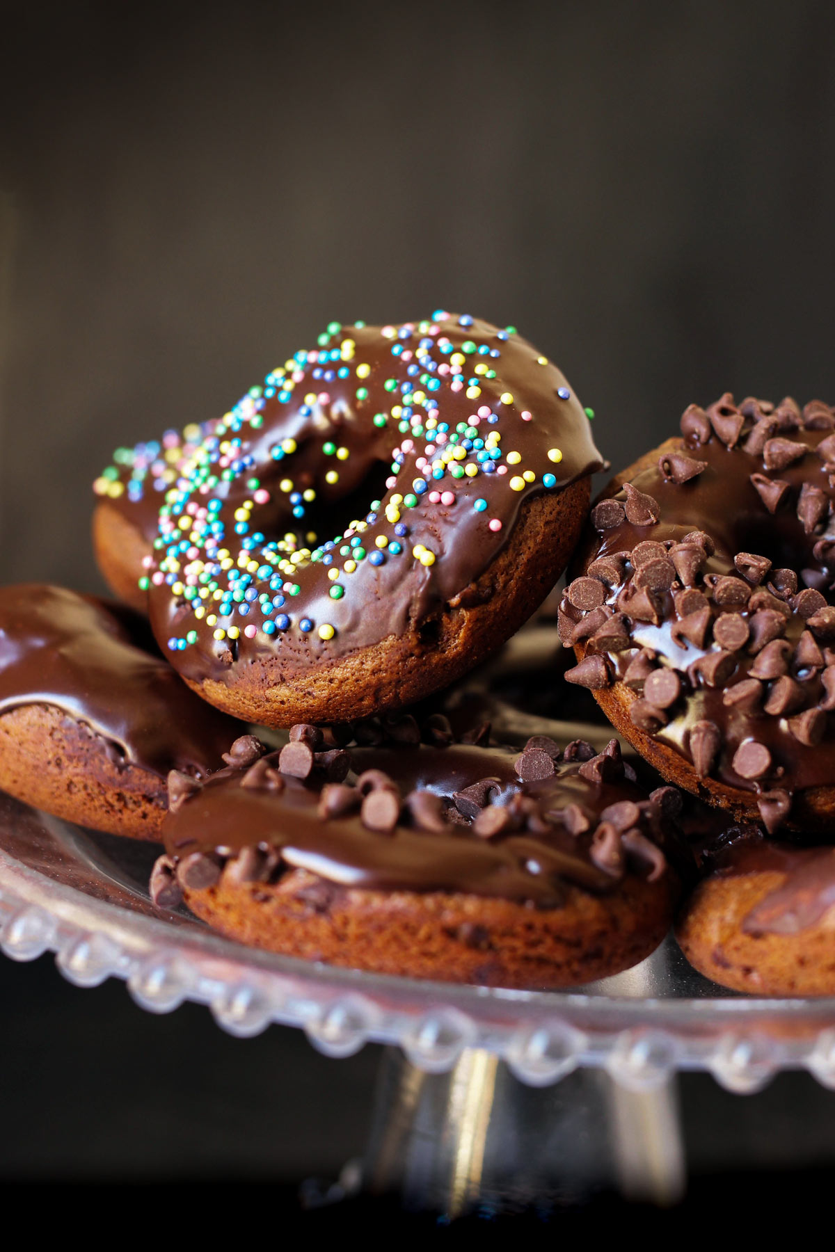 stack of chocolate baked donuts with different sprinkles and mini chips on cake stand