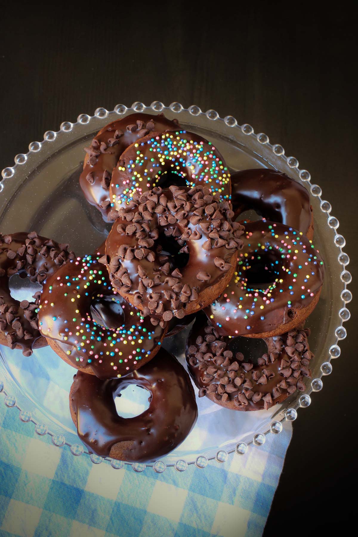 overhead shot of a stack of chocolate donuts on a cake stand with a blue check napkin.