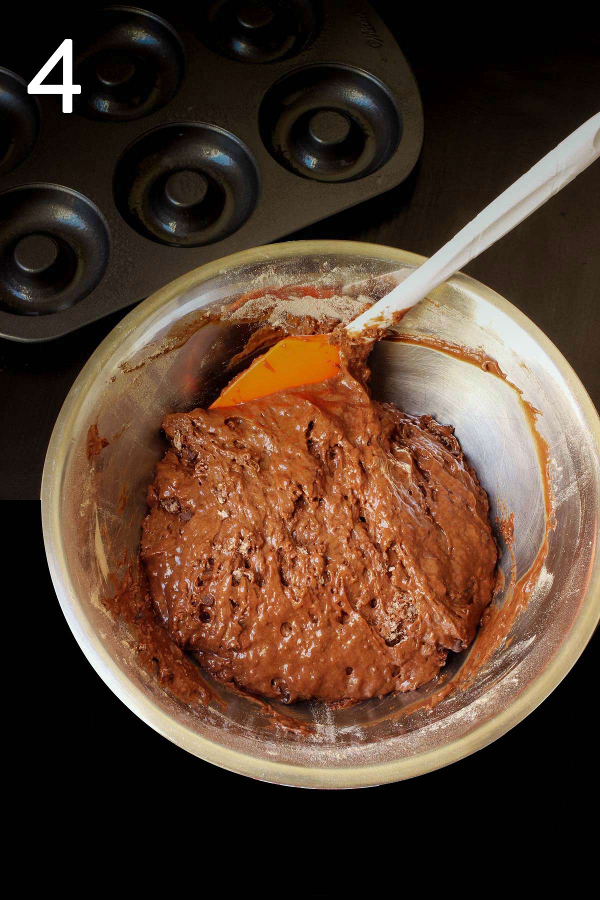 the chocolate donut batter assembled in a metal bowl with an orange spatula.
