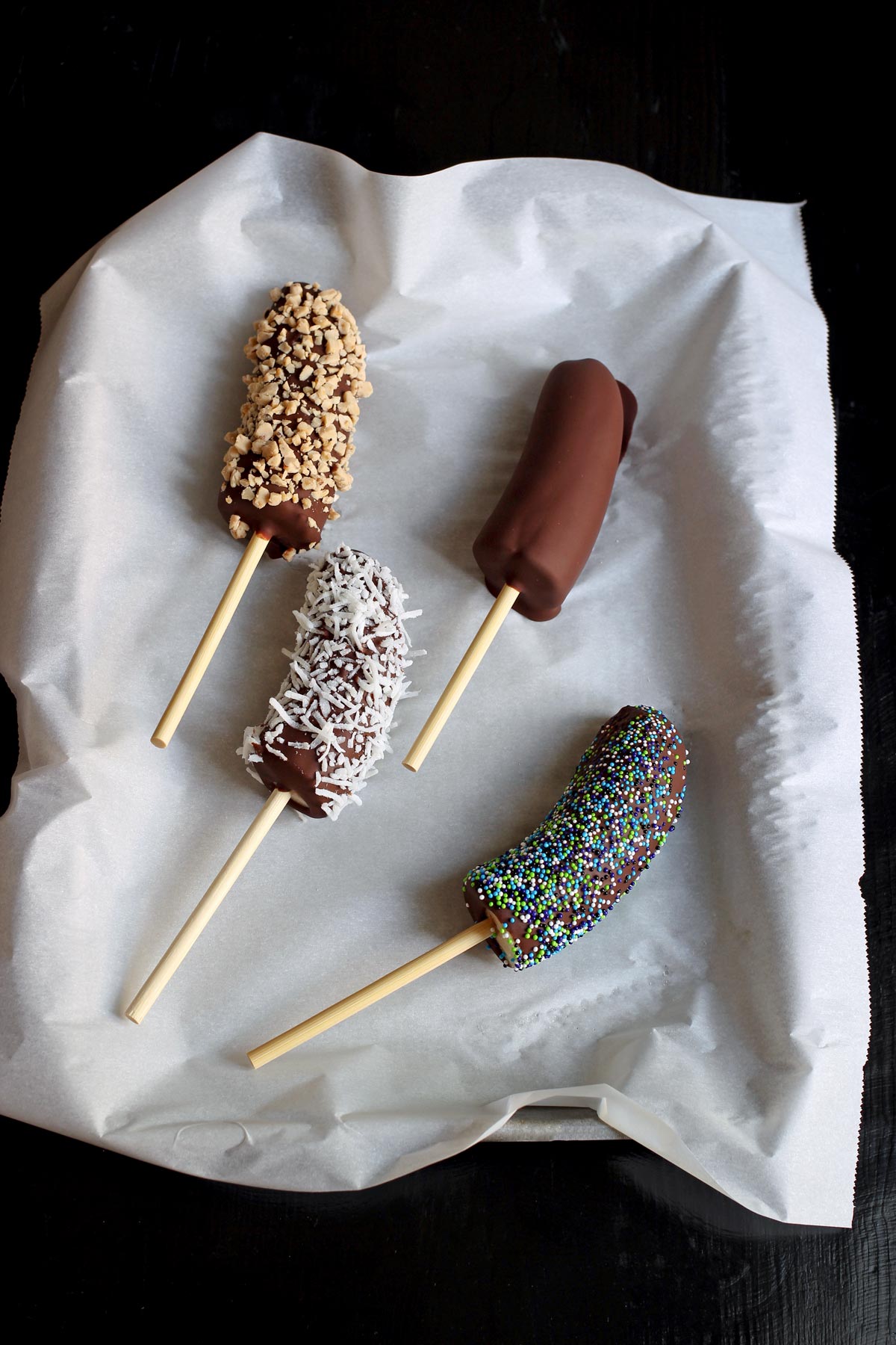 four frozen bananas dipped in chocolate with four different toppings on a tray lined with parchment.