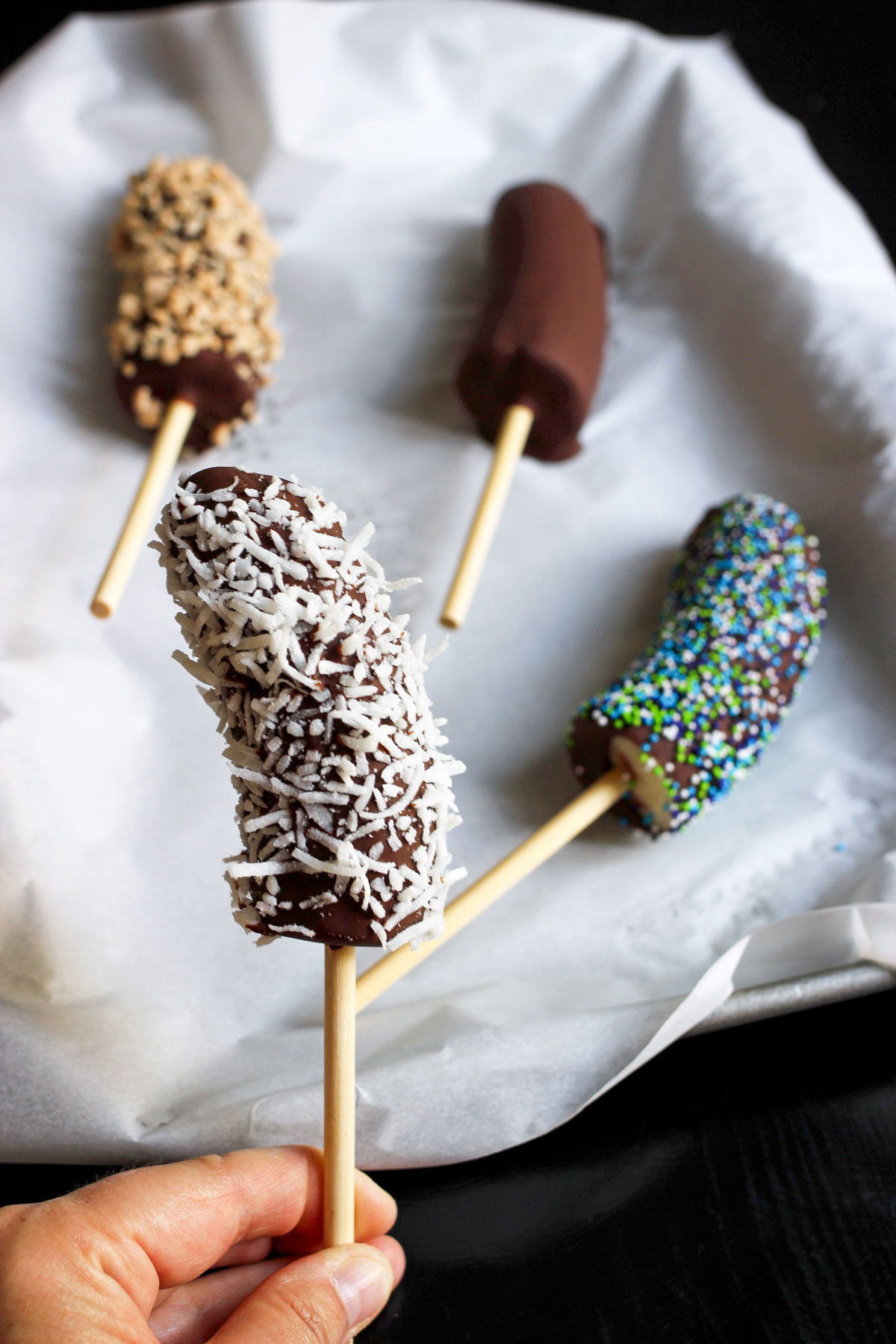 hand holding chocolate dipped banana coated with coconut in front of three frozen bananas with other toppings on a tray.