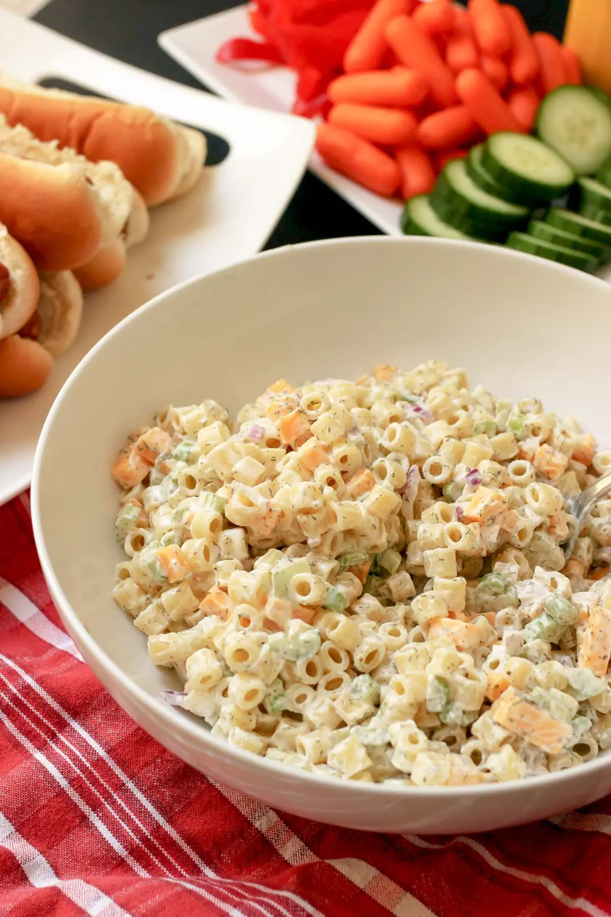 white bowl of macaroni salad on red cloth near platters of hot dogs and veggies.