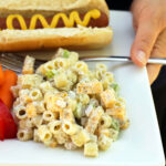 hand holding square plate with macaroni salad, fruit salad, and a hot dog with mustard.
