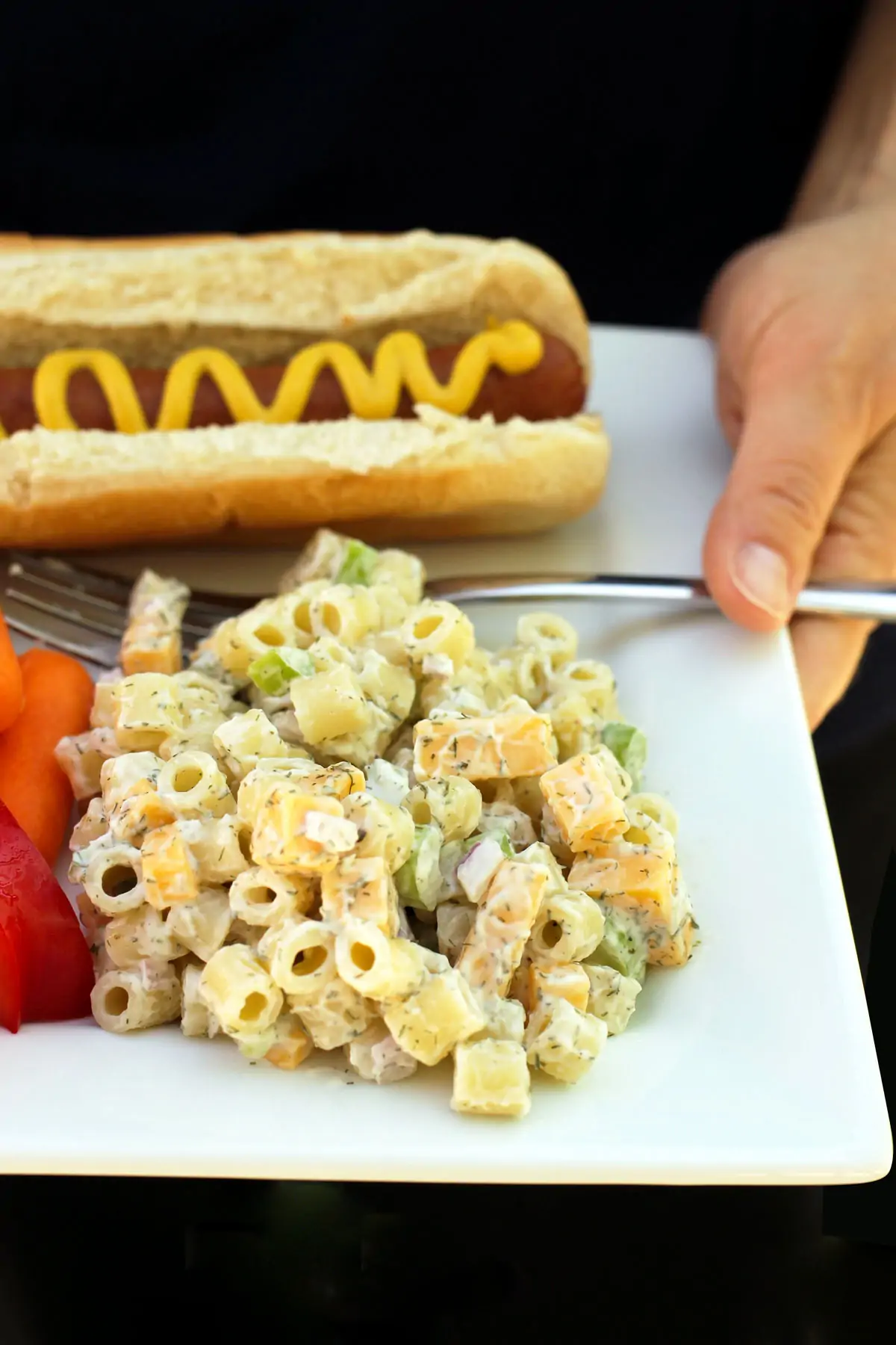 woman holding white, square plate of macaroni salad, veggies, and a hot dog with mustard.