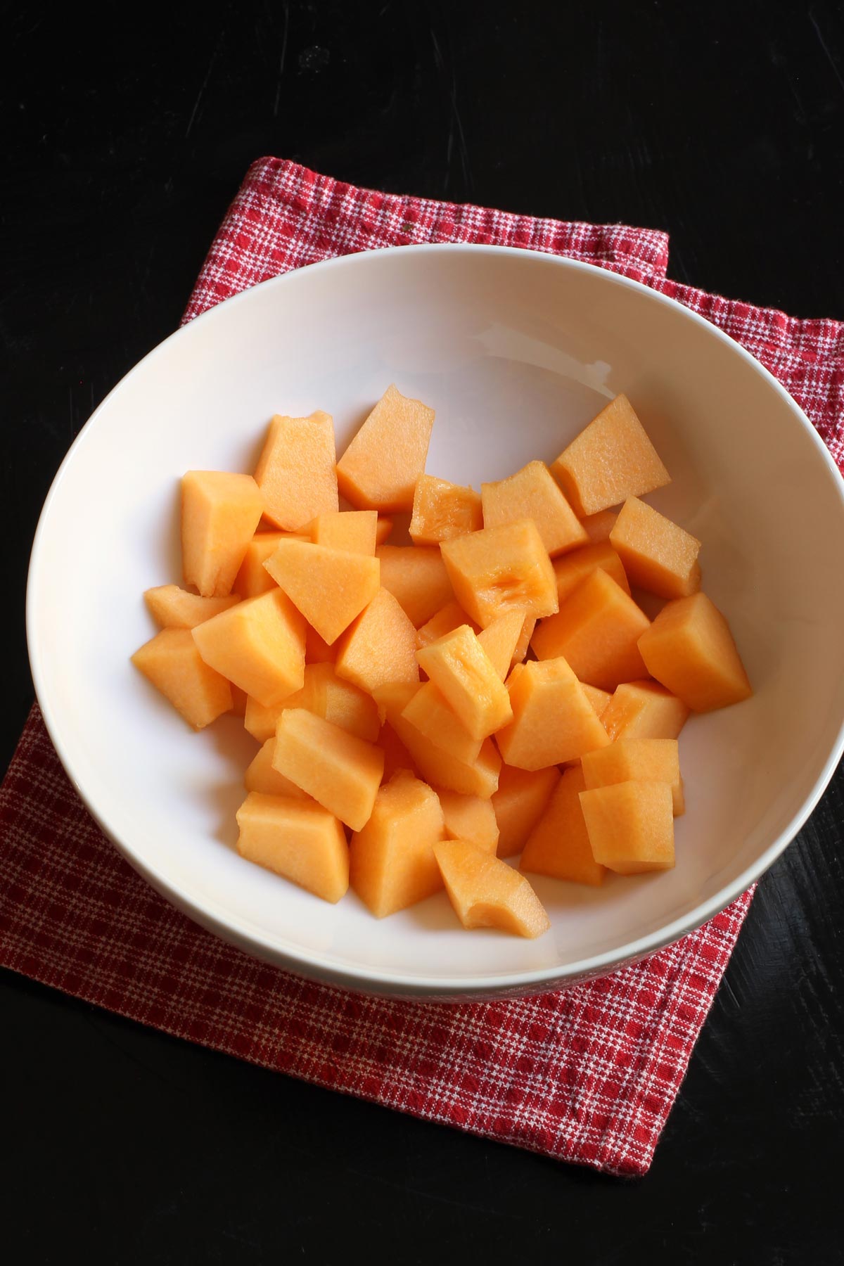 large white bowl of melon cubes atop a red checked cloth.