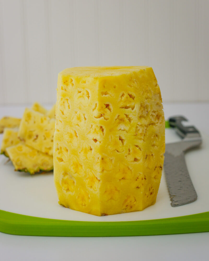 the trimmed pineapple standing upright on the cutting board with skin in the background. 