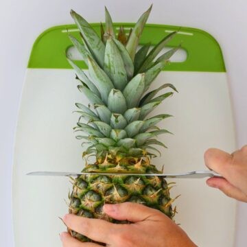 hands holding a pineapple on a cutting board to cut off the top with a knife.