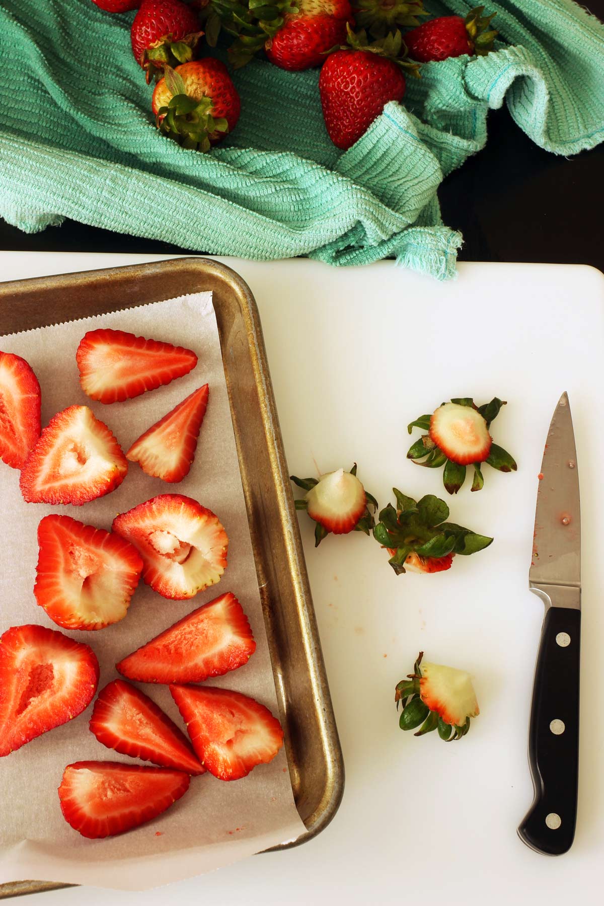 strawberry cores and knife on cutting board next to tray of strawberries prepped for freezing.