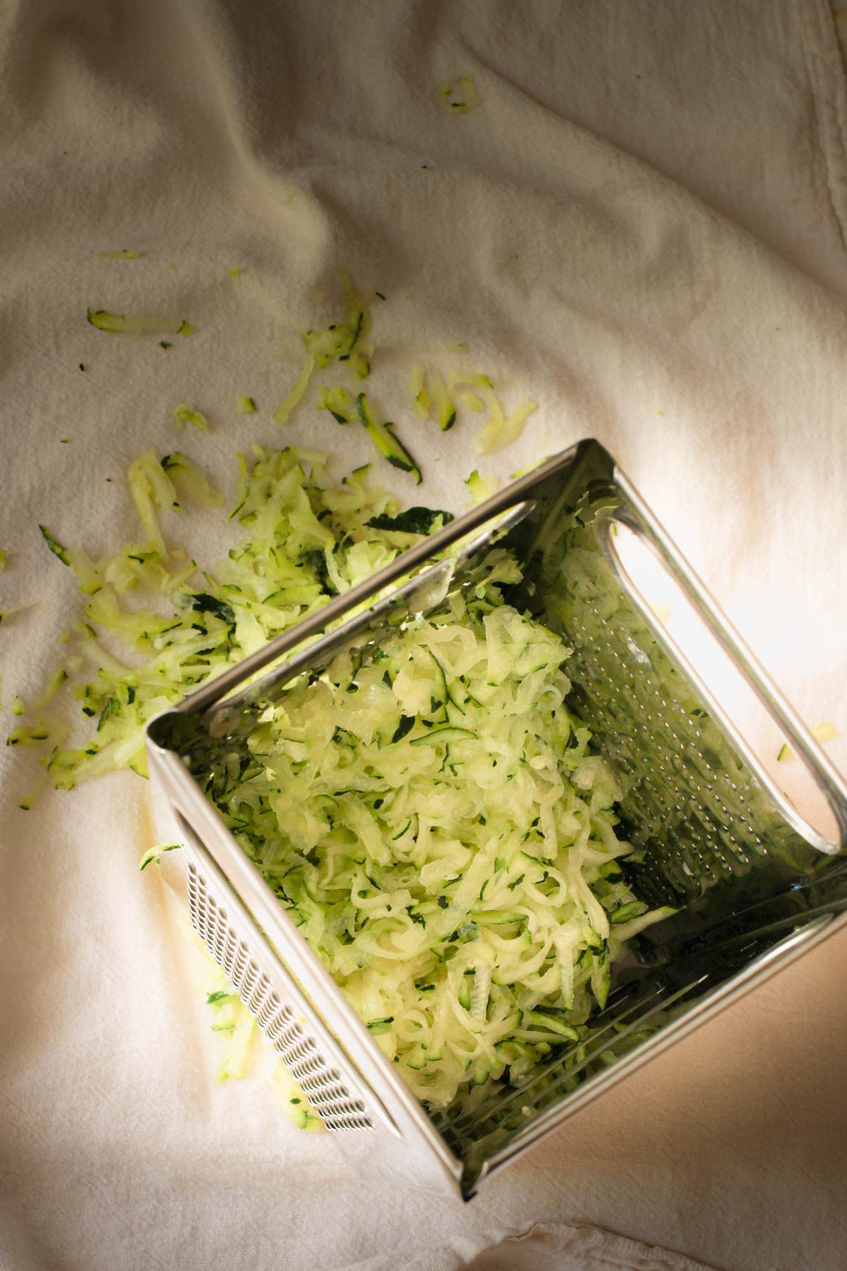 box grater on white tea towel with shredded zucchini.