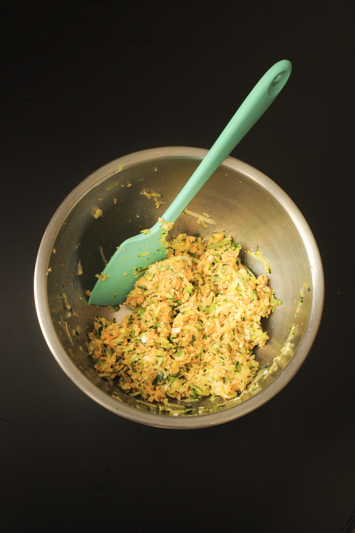 zucchini fritter mixture combined in mixing bowl with teal spatula.
