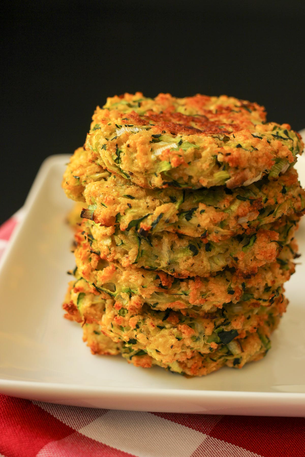 stack of baked zucchini fritters on a white square plate on red-checked napkin.