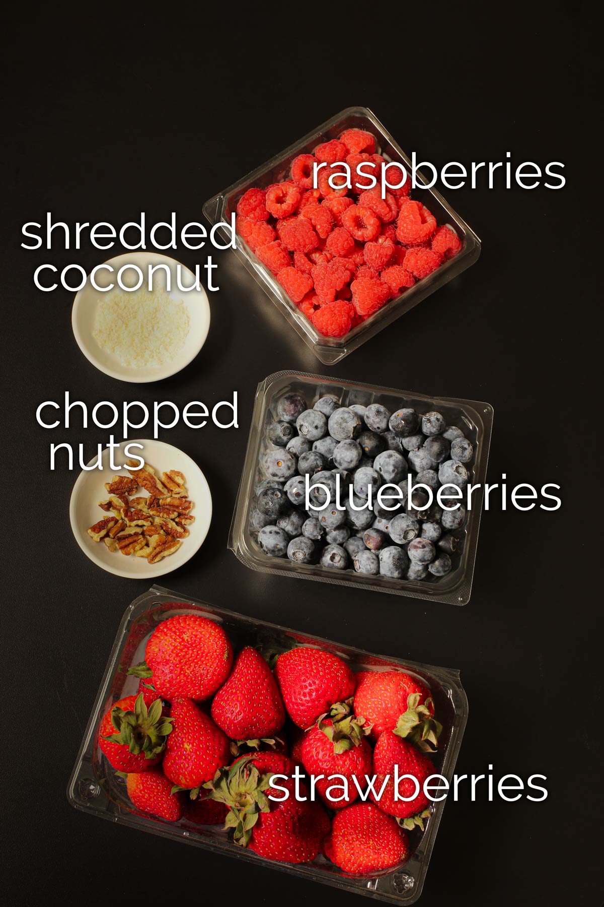 cartons of berries next to dishes of nuts and coconut on black tabletop.