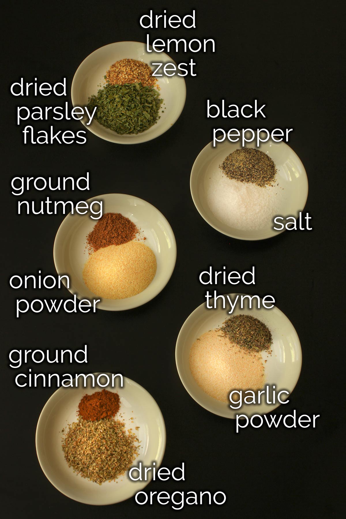 ingredients for Greek spice blend measured into small white bowls on black tabletop.