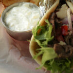 metal cup of garlic yogurt sauce next to greek pita sandwich in lined basket.