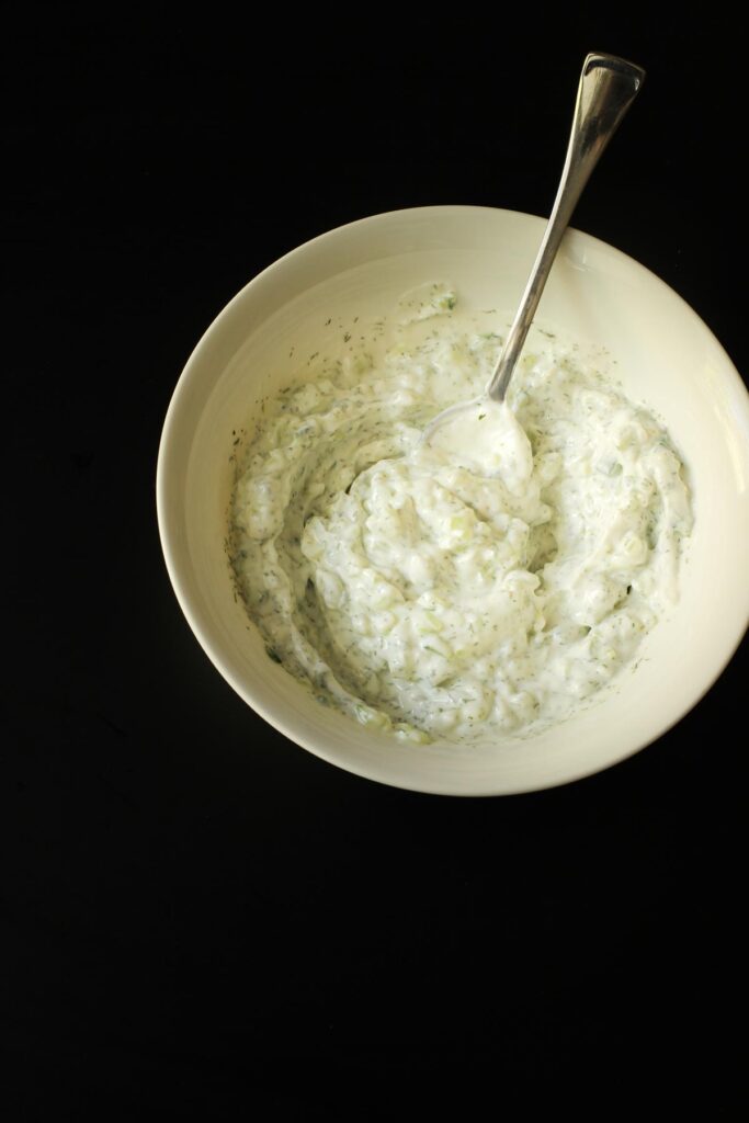 white bowl of garlic yogurt sauce with a silver spoon in the swirls.