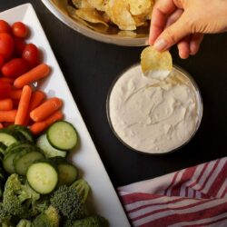 hand dipping a potato chip into bowl of sour cream onion dip near platter of veggie dippers.