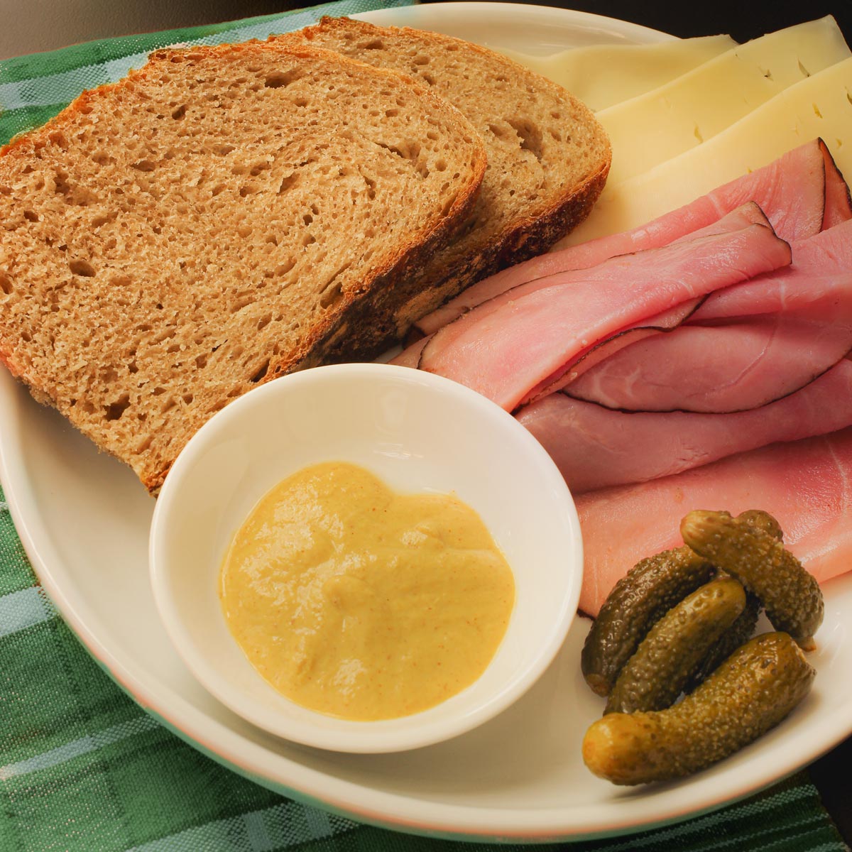 plate of sourdough rye bread slices with ham, cheese, pickles, and mustard.