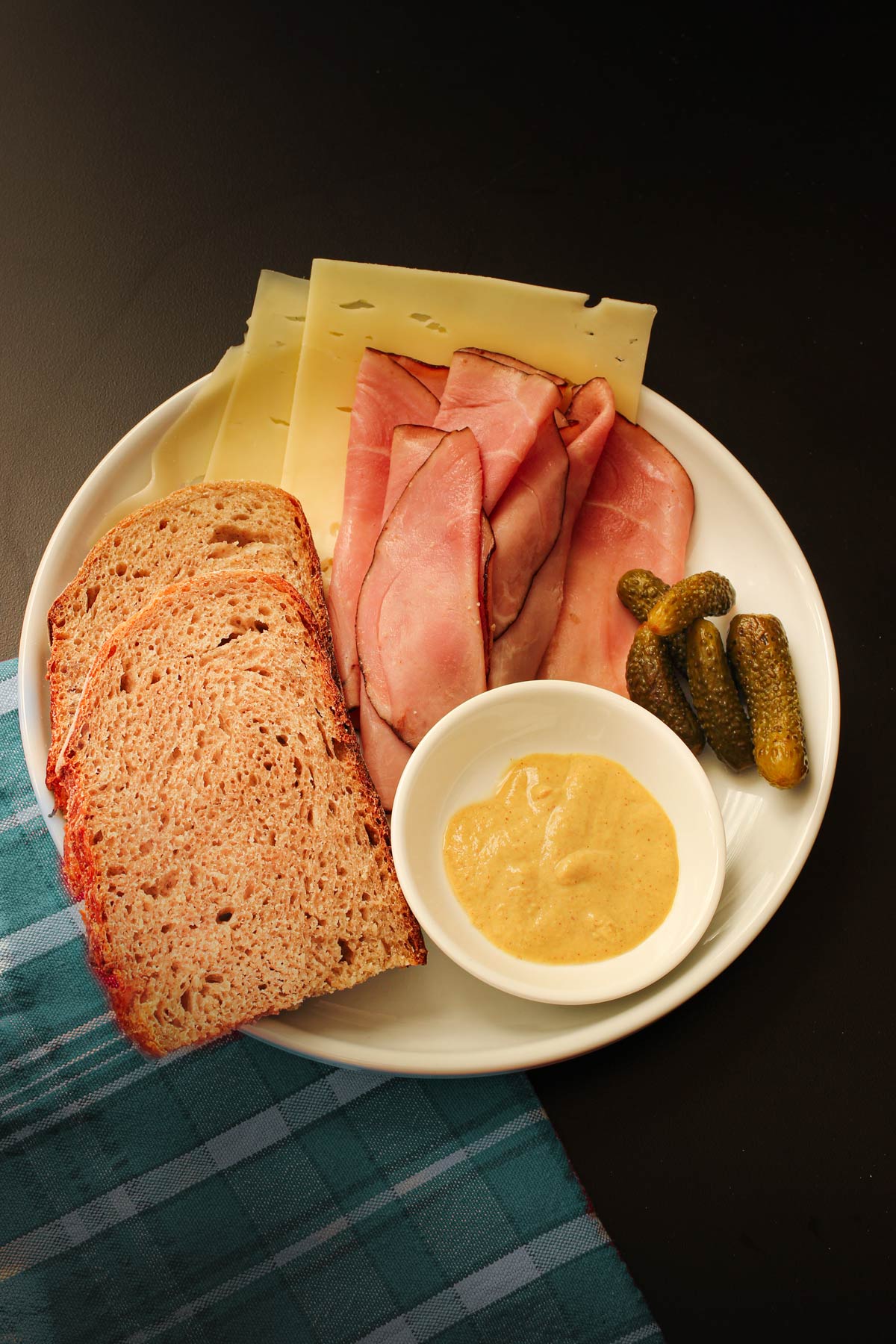 plate with slices of sourdough rye bread as well as ham, cheese, cornichons, and a small dish of mustard.