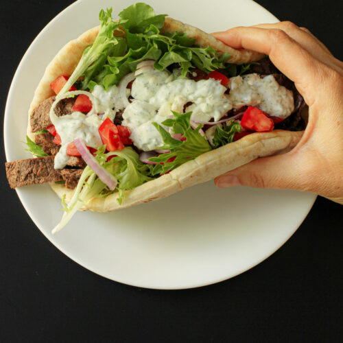 hand holding unwrapped steak pita sandwich with all the toppings.