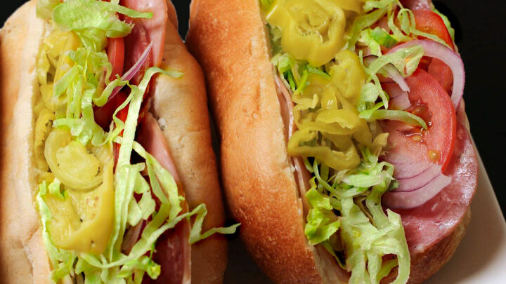 Italian sub sandwiches on platter bursting with cold cuts and other fillings.