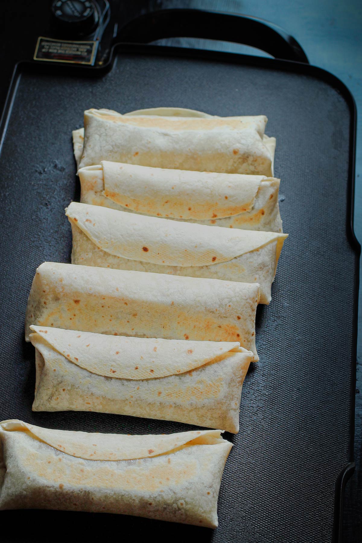 six beef chimichangas crisping on a large electric griddle.
