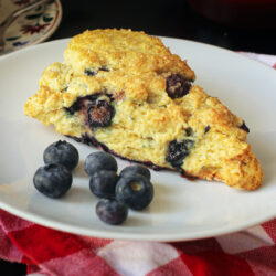 close up of a lemon blueberry scone on a white plate with a handful of fresh blueberries.