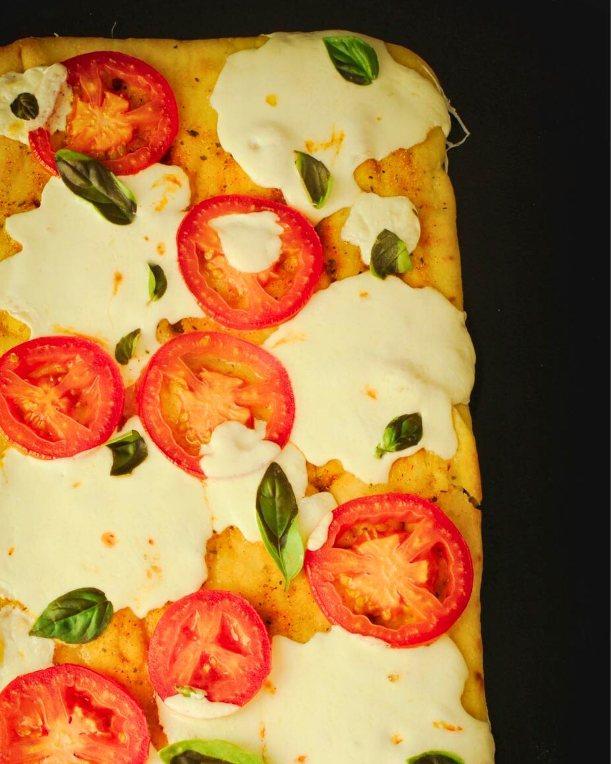 close up of baked margherita flatbread on a black background.