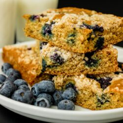 stacked squares of blueberry lemon coffeecake on a plate with blueberries with glasses of milk in background.