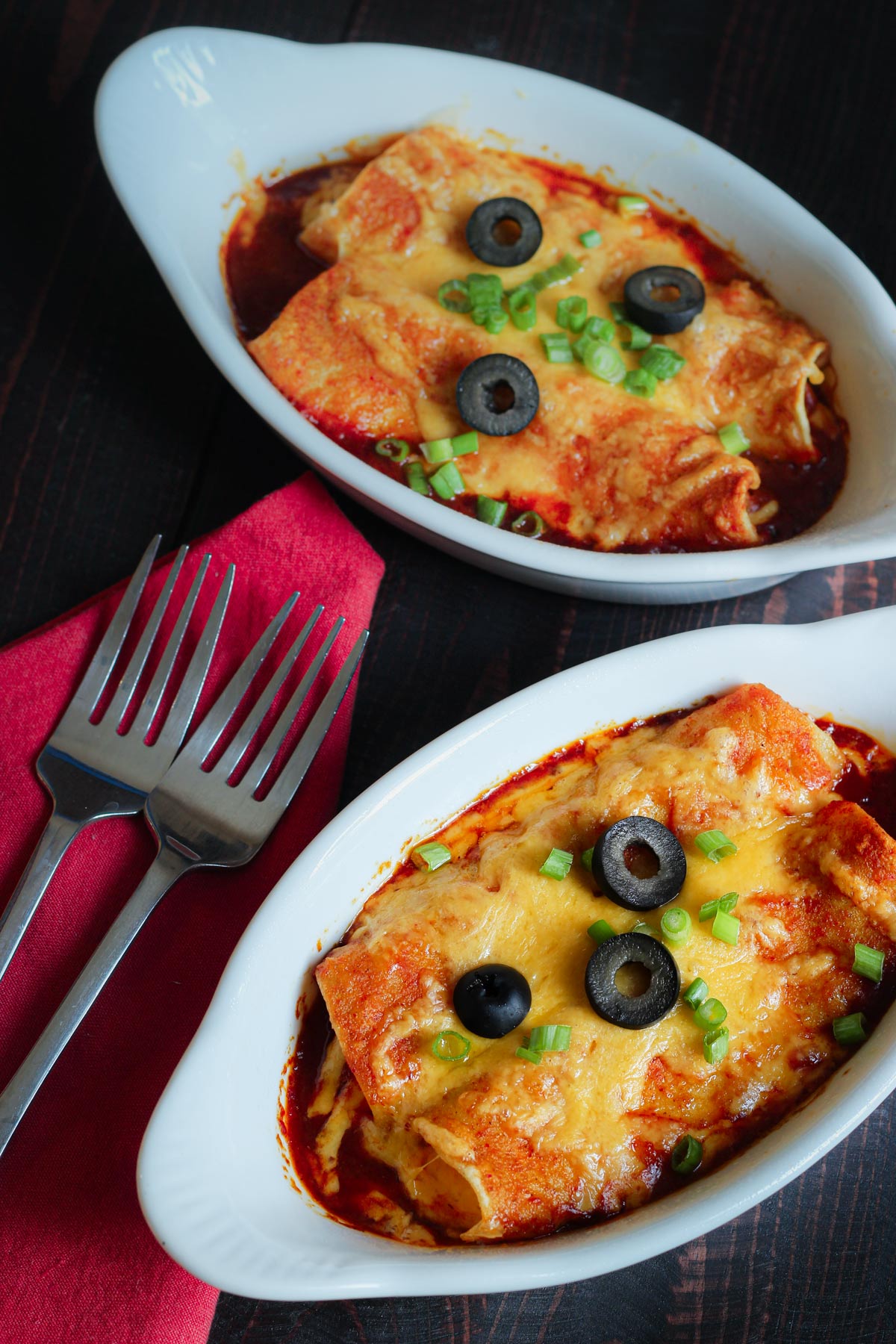 pair of gratin dishes filled with cheese enchiladas next to red napkin with two forks.