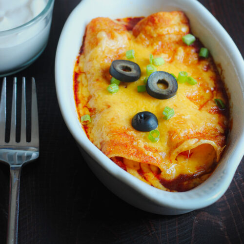white gratin dish with two cheese enchiladas topped with olives and green onions next to fork and dish of sour cream on the table.