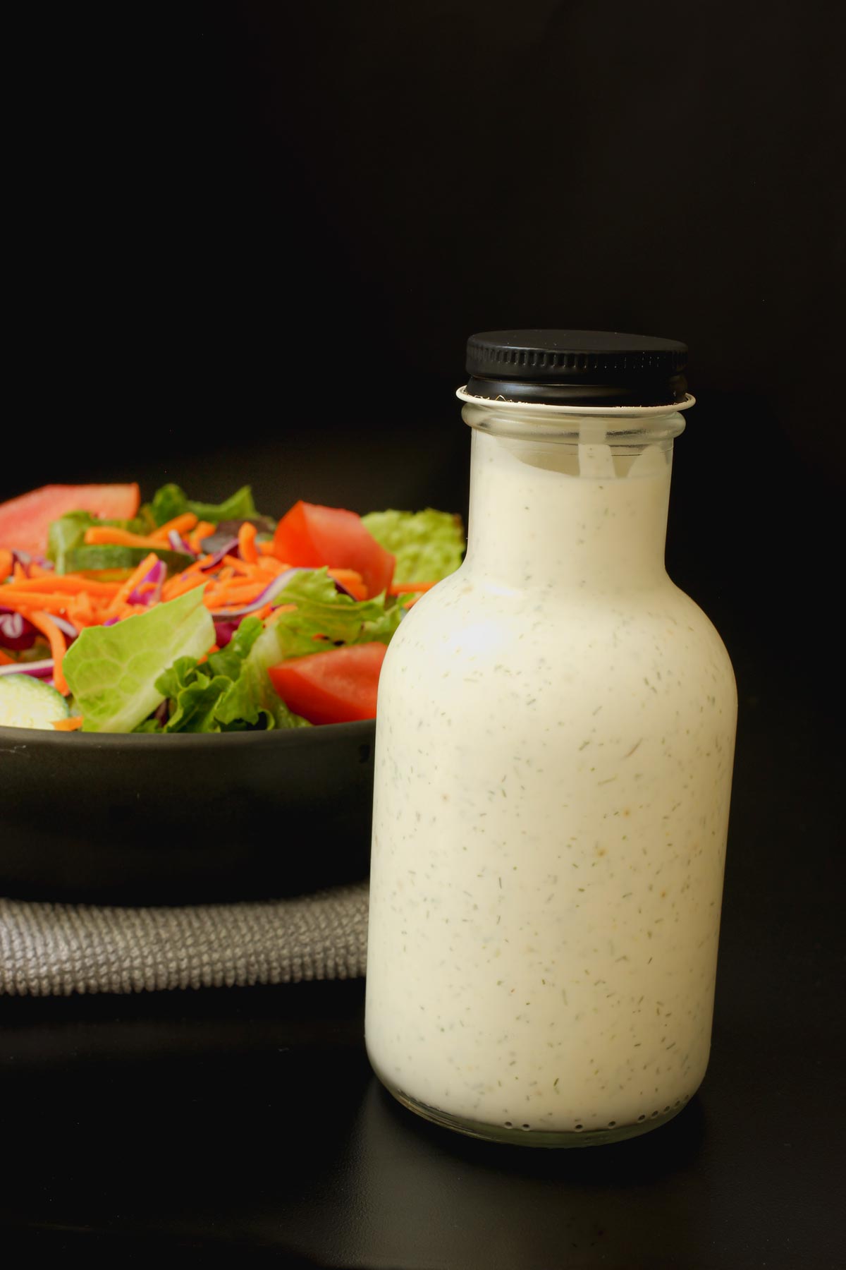 closed bottle of buttermilk dressing next to salad in a black bowl.