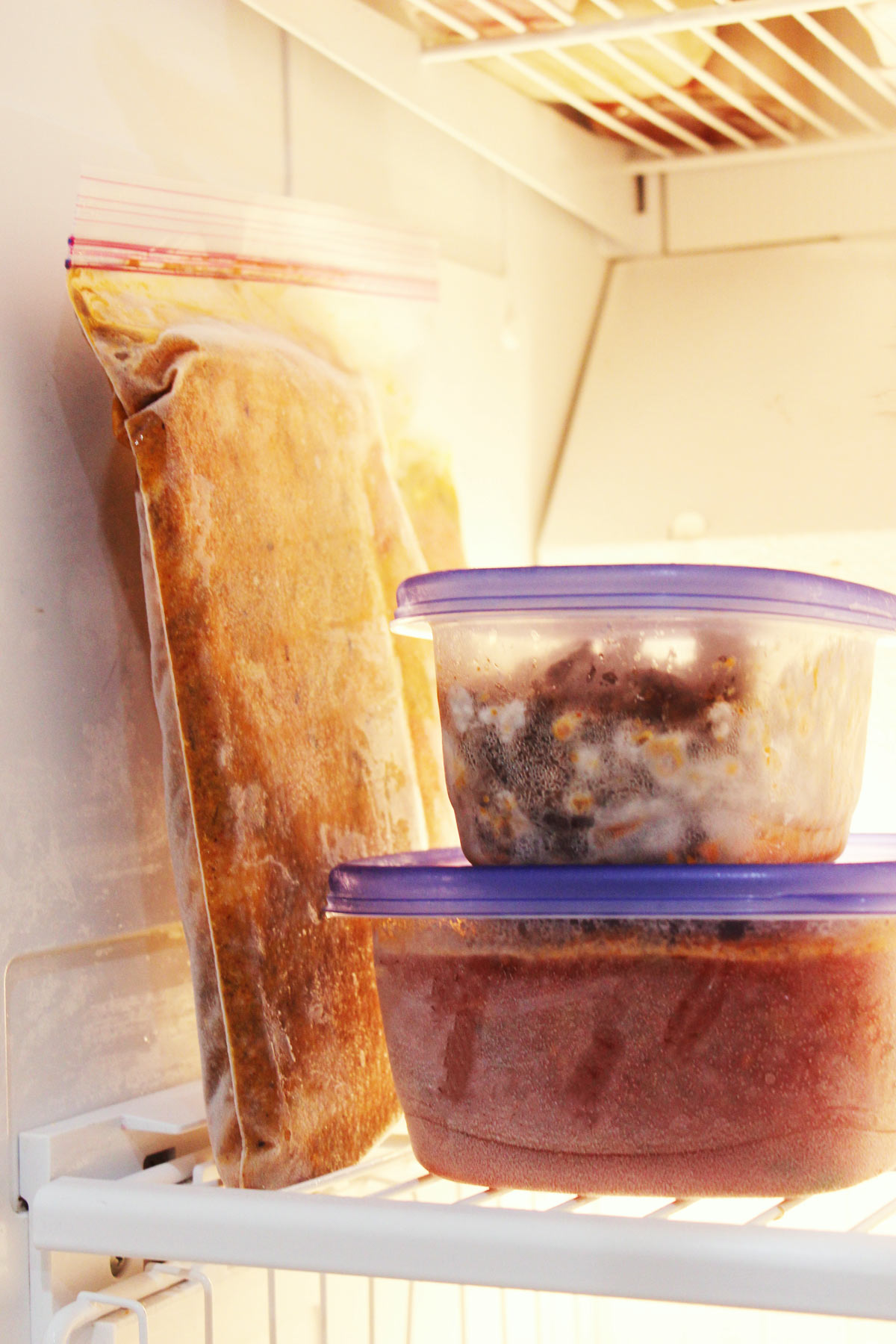 bagged freezer meal leaning against side of freezer wall next to stacked plastic containers of food on freezer shelf.