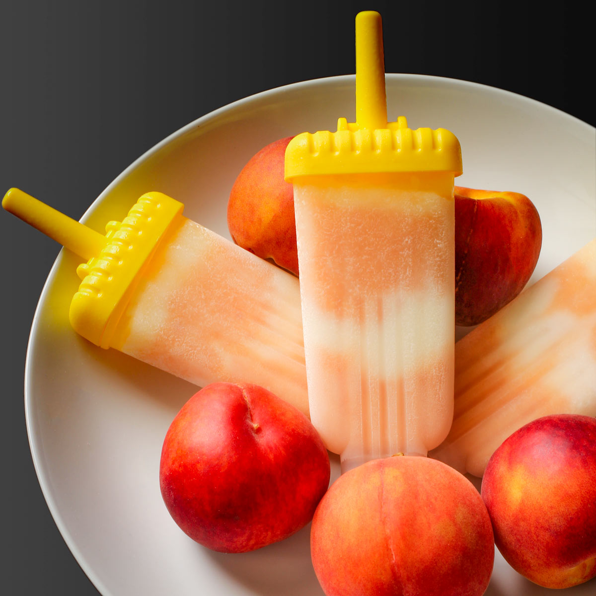 peach yogurt popsicles in their molds in a large white bowl of peaches.