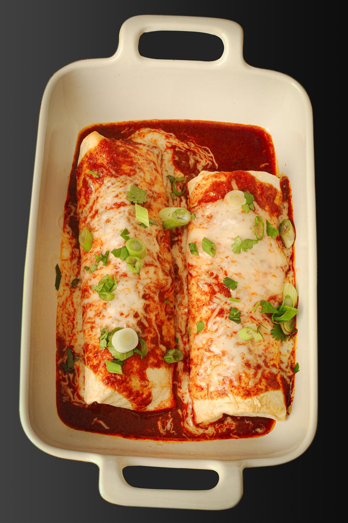 two chimichangas in a baking dish, topped with sauce, cheese, and green onions.