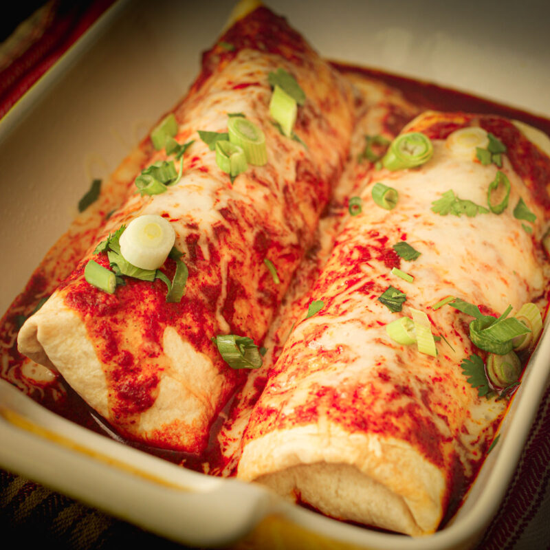 pair of chimichangas in baking dish topped with sauce, cheese, and chopped scallions.