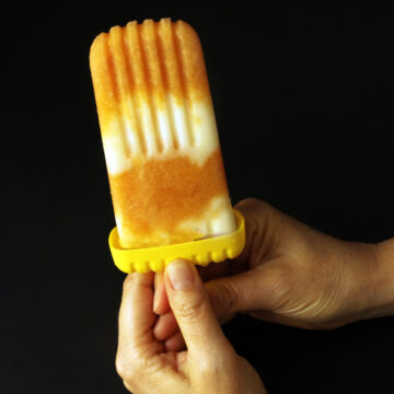 hands holding peach yogurt popsicle against black backdrop.