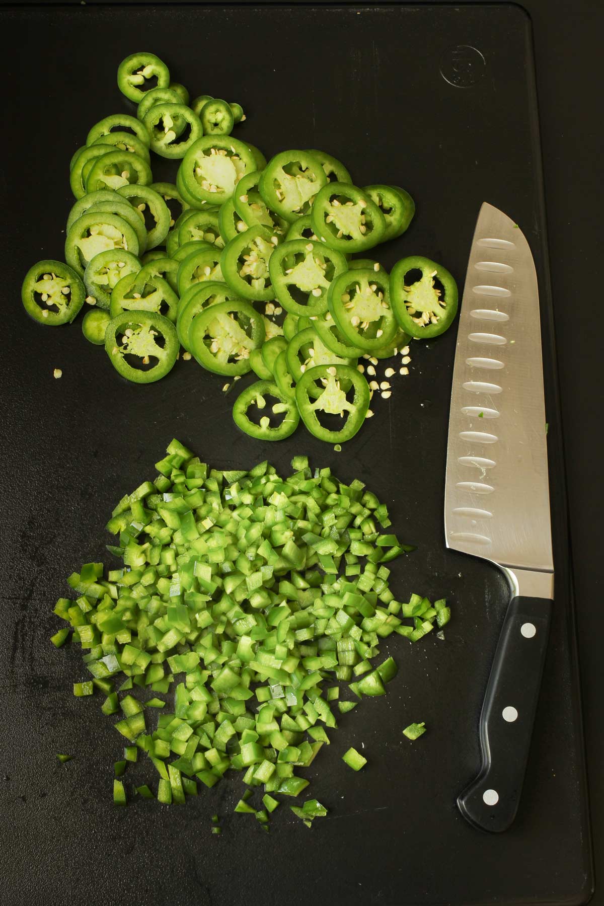 sliced and chopped jalapeños on black cutting board with knife nearby.