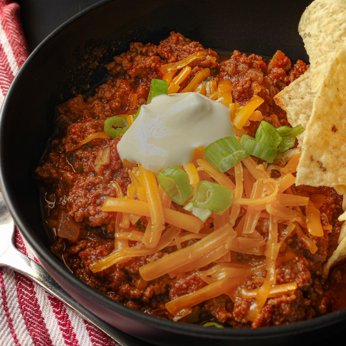 no bean chili recipe served in a black bowl topped with cheese, sour cream, green onions, and chips.