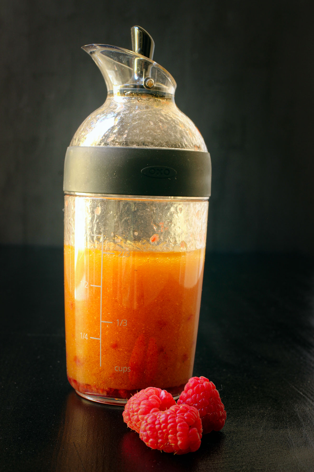 raspberry vinaigrette in cruet on black table with fresh raspberries nearby.