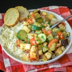 ratatouille on a bed of rice on a white plate with slices of baguette.