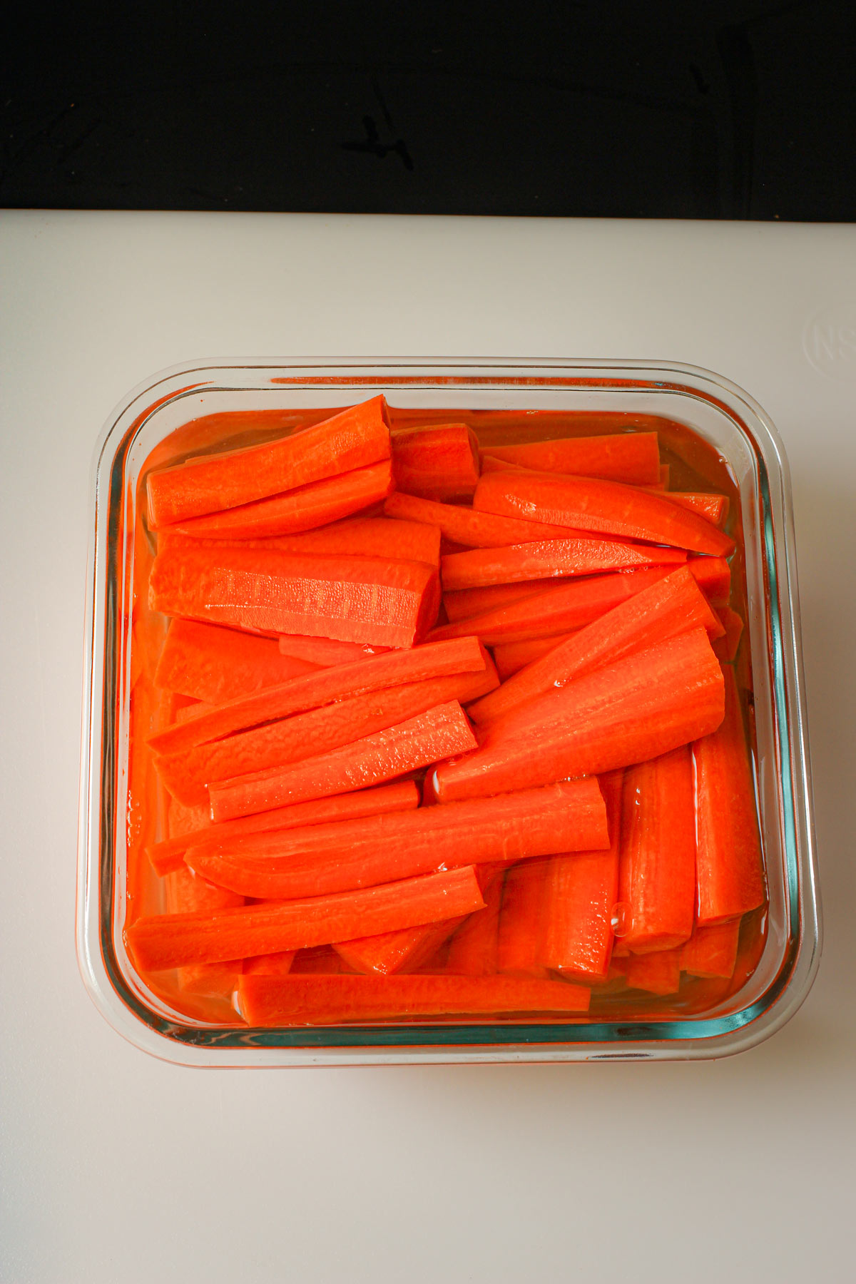 carrot sticks floating in water in glass storage container on white cutting board.