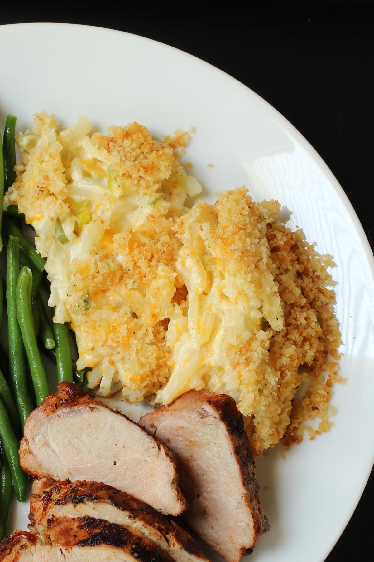dinner plate closeup view of cheese hashbrown casserole with green beans and grilled pork.
